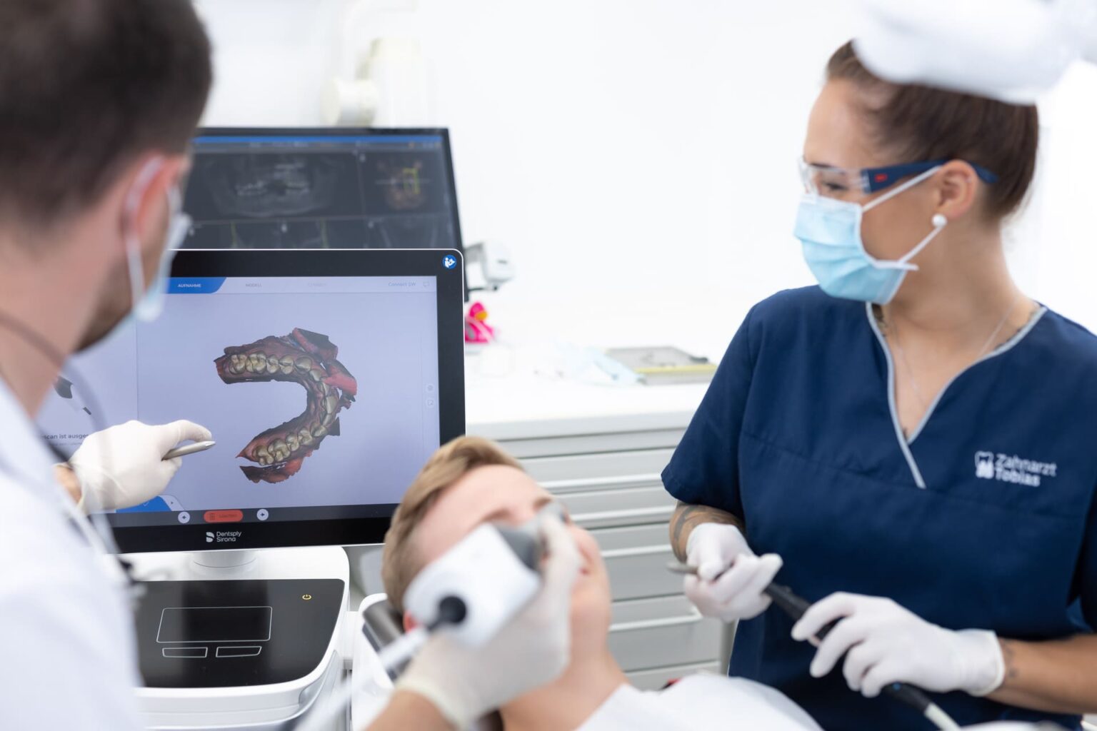 Zahnarzt und Assistenz in blauer Arbeitskleidung behandeln Patient im Behandlungsstuhl, Zahnbild auf Monitor sichtbar.