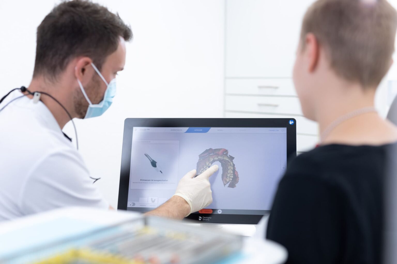 Zahnarzt zeigt mit Handschuh auf 3D-Darstellung eines Kiefers auf Bildschirm, Patient sitzt daneben.